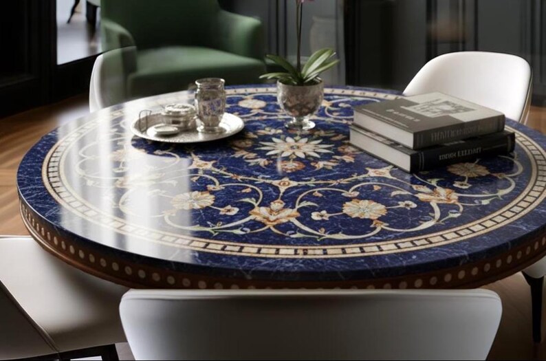 Pu&ograve; includere: Un tavolo rotondo con una superficie blu scuro, ornato da un motivo floreale color crema e oro. Il tavolo ha un bordo in legno ed &egrave; circondato da sedie bianche. Oggetti decorativi e libri sono posizionati sul tavolo.