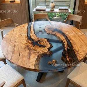Puede incluir: Una mesa de madera grande y redonda con un diseño único. La mesa presenta una veta de madera marrón claro con detalles de resina marrón oscuro y azul. Se apoya en una base de metal negro y está rodeada de sillas de madera.