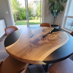 Puede incluir: Una mesa de comedor redonda con un centro de vetas de madera natural y un borde de resina negra. La mesa está rodeada de sillas de cuero marrón y está situada en un comedor luminoso con grandes ventanas.