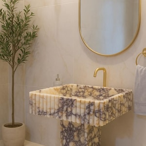 Puede incluir: Un lavabo de mármol con una cubeta rectangular y un borde acanalado, sostenido por un pedestal a juego. El lavabo tiene un grifo dorado y está colocado contra una pared alicatada de tonos neutros. También se ven un espejo con marco dorado y una pequeña planta en maceta.