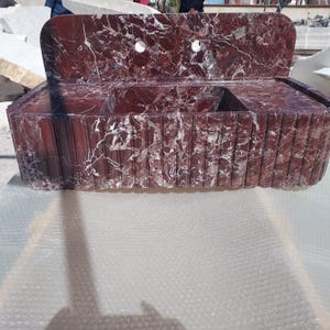May include: A red marble sink with white veining. The sink has a rectangular basin with a raised back and two circular faucet holes. The front of the sink has a vertical grooved design.