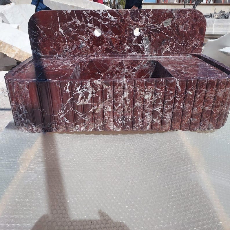 May include: A red marble sink with white veining. The sink has a rectangular basin with a raised back and two circular faucet holes. The front of the sink has a vertical grooved design.