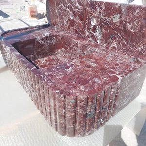May include: A red marble bathroom sink with a built-in backsplash. The sink has a rectangular basin and a decorative, fluted front. The marble features a striking pattern of red and white veining, creating a luxurious aesthetic.
