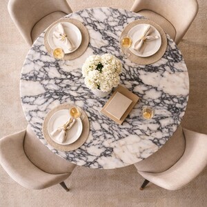 May include: Overhead view of a round marble dining table with four beige upholstered chairs. The table is set with plates, napkins, and glasses, and a vase of white flowers sits in the center.