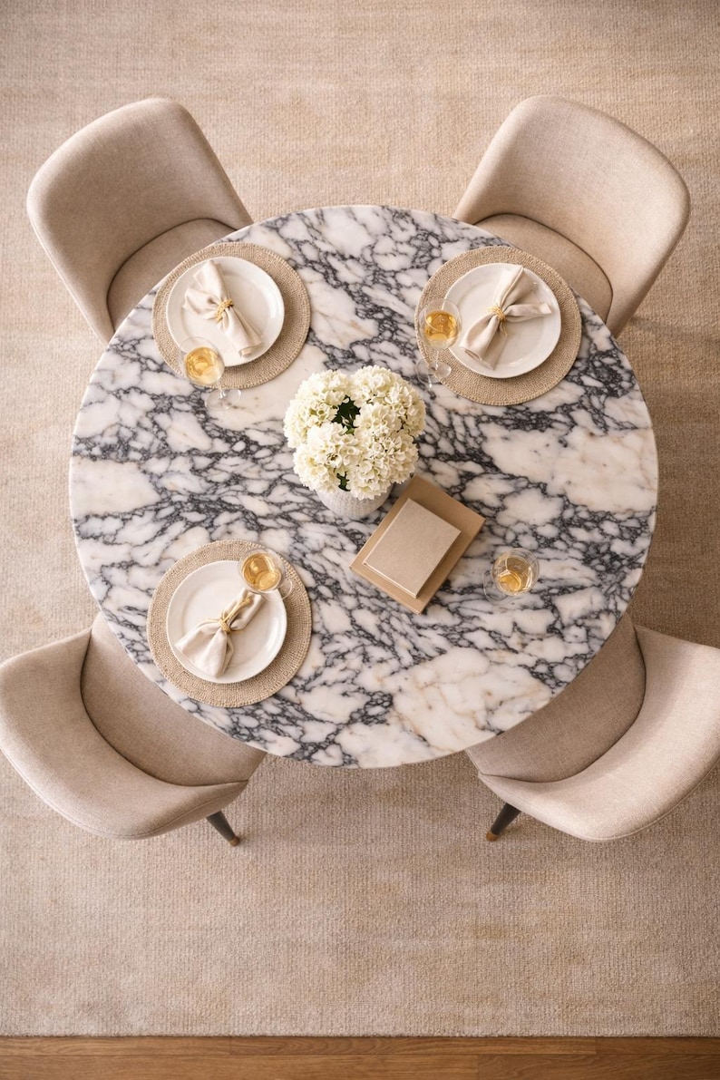 May include: Overhead view of a round marble dining table with four beige upholstered chairs. The table is set with plates, napkins, and glasses, and a vase of white flowers sits in the center.