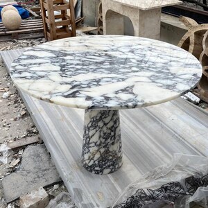 May include: A round marble table with a white and gray patterned top and a cylindrical base. The table is made of stone and is suitable for indoor or outdoor use. The table's diameter is approximately 48 inches.