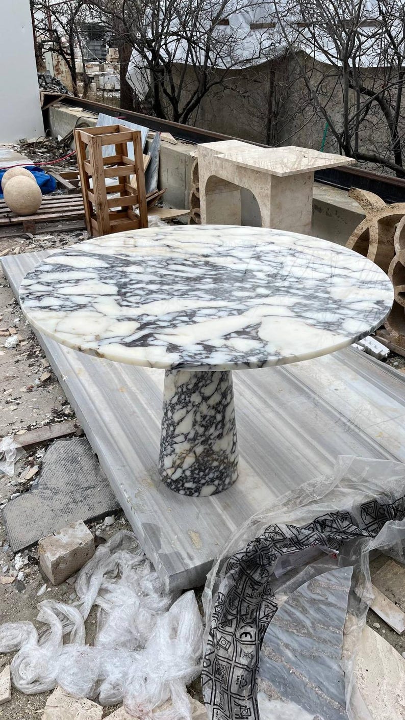 May include: A round marble table with a white and gray patterned top and a cylindrical base. The table is made of stone and is suitable for indoor or outdoor use. The table's diameter is approximately 48 inches.