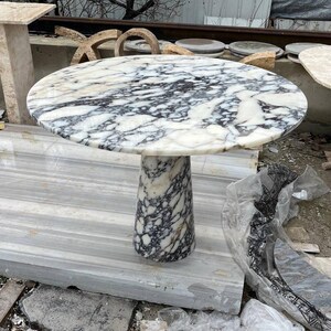 May include: A round marble table with a white and black veined pattern. The table has a cylindrical base and is approximately 30 inches in diameter. The table is made of stone and is suitable for indoor or outdoor use.
