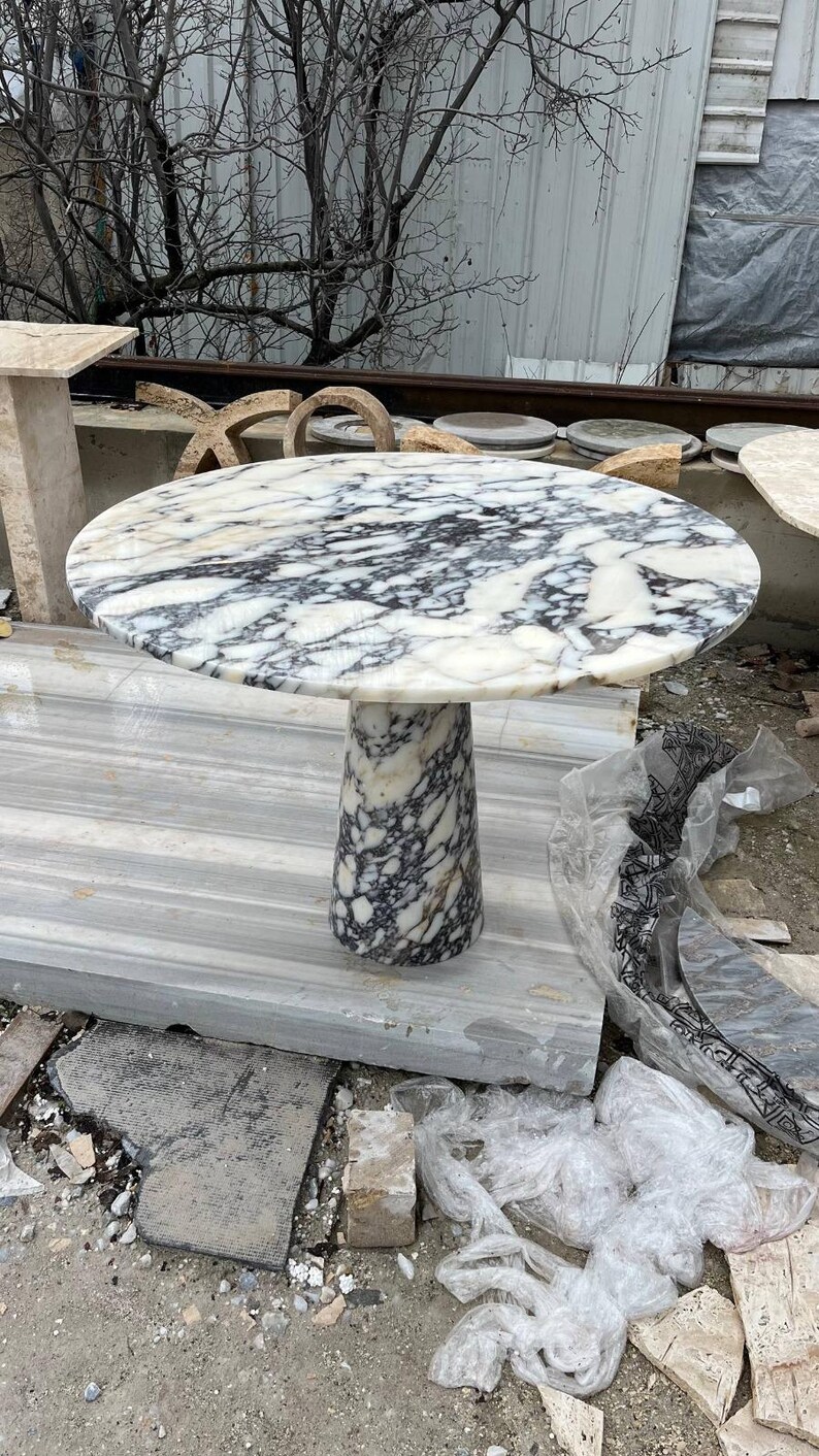 May include: A round marble table with a white and black veined pattern. The table has a cylindrical base and is approximately 30 inches in diameter. The table is made of stone and is suitable for indoor or outdoor use.
