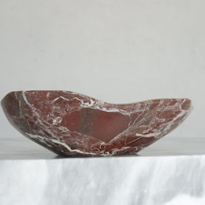 May include: A red marble bowl with white veining. The bowl has an organic shape and sits on a white surface. The interior of the bowl is a darker shade of red, creating a contrast with the exterior.