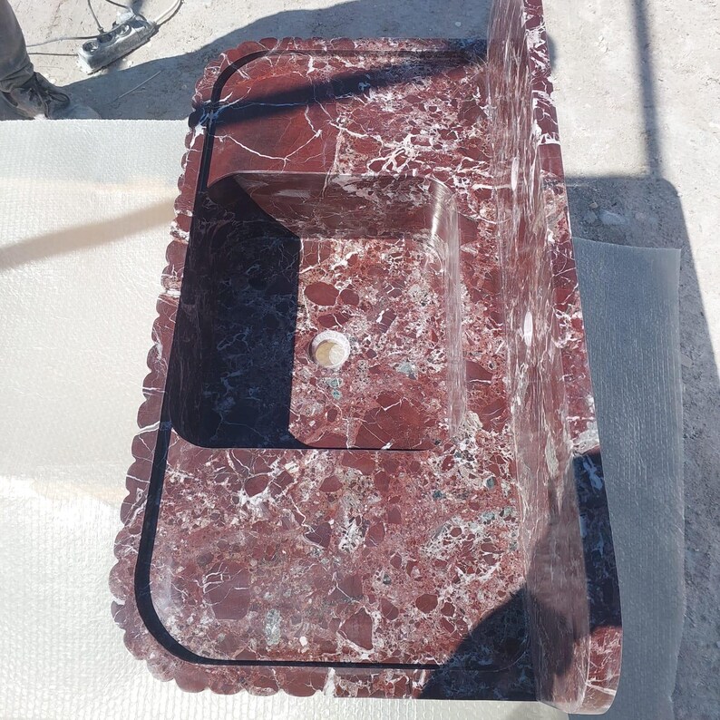 May include: A red marble sink with a rectangular basin and a raised backsplash. The sink has a scalloped edge and a speckled pattern of white and dark red. The faucet holes are visible.