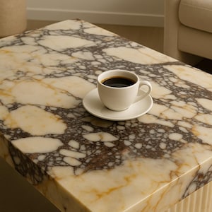 Puede incluir: Una mesa de centro rectangular con una tapa de mármol con un patrón crema, marrón y dorado. Una taza y un platillo blancos con café negro están sobre la mesa. La base de la mesa tiene un diseño acanalado vertical.