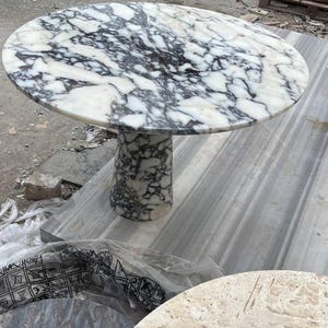 May include: A round marble table with a white and black patterned top. The table has a cylindrical base and is likely used for dining or as a decorative piece. The table's diameter is approximately 48 inches.