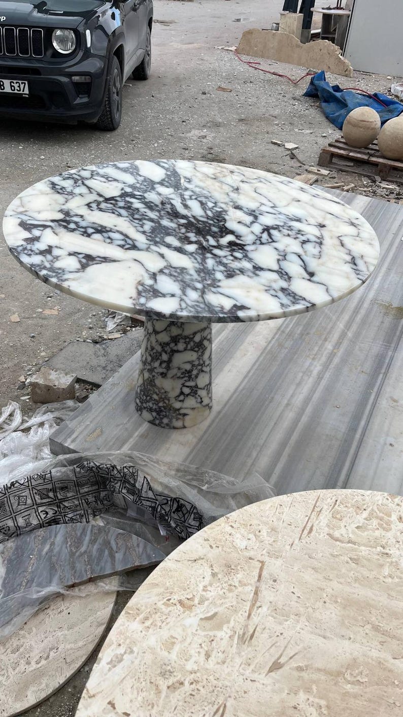 May include: A round marble table with a white and black patterned top. The table has a cylindrical base and is likely used for dining or as a decorative piece. The table's diameter is approximately 48 inches.