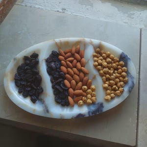 Könnte beinhalten: Eine ovale Servierplatte mit drei Abschnitten, die jeweils einen Snack enthalten. Die Platte zeigt dunkle Rosinen, Mandeln und geröstete Kichererbsen. Das Gericht hat ein marmoriertes Aussehen mit weißen, beigen und braunen Farbtönen.
