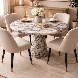 May include: A round marble dining table with a gray and white pattern, supported by a matching pedestal base. Four beige upholstered chairs with black legs and gold tips surround the table. The setting includes a rug, flowers, and wine glasses.
