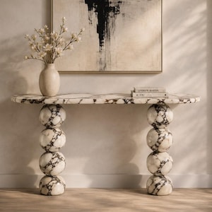 May include: A marble console table with a patterned top and spherical legs. The table supports a vase with white flowers and a stack of books. An abstract black and white painting hangs on the wall behind the table.