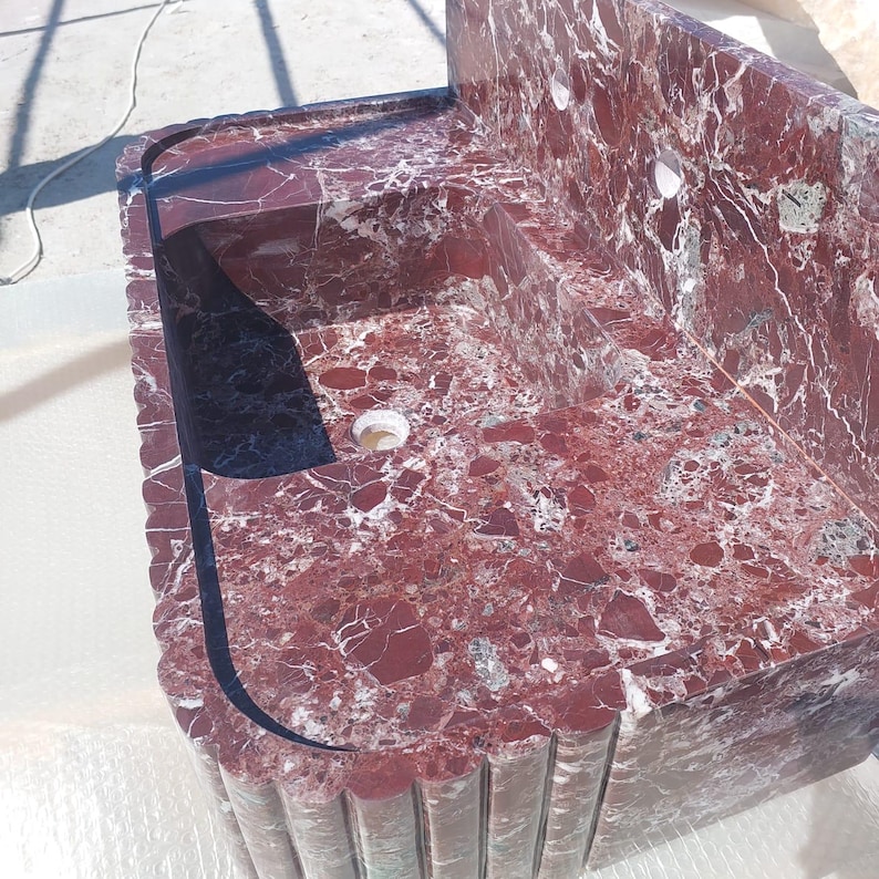 May include: A red marble sink with white veining. The sink has a rectangular basin with a curved edge and a built-in backsplash. The sink has a drain hole and is made of a polished stone material.