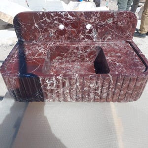 May include: A red marble sink with white veining. The sink has a rectangular basin with a raised back and two faucet holes. The front of the sink has a fluted design. A modern bathroom fixture.