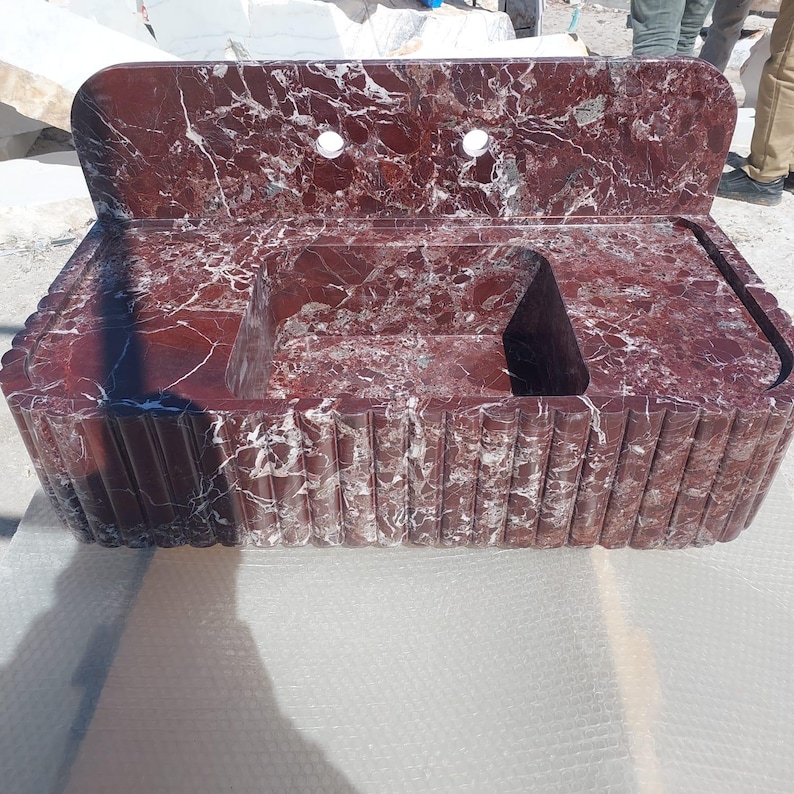 May include: A red marble sink with white veining. The sink has a rectangular basin with a raised back and two faucet holes. The front of the sink has a fluted design. A modern bathroom fixture.