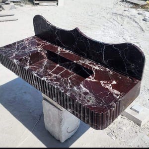 Op de afbeelding: Een rechthoekige, donkerrode marmeren wastafel met witte adering. De voorrand heeft een groefontwerp en een gebogen backsplash komt van de achterkant. De wastafel wordt ondersteund door een witte stenen basis.