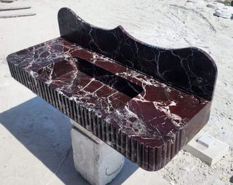 Puede incluir: Un tocador rectangular de mármol rojo oscuro con vetas blancas. El borde frontal tiene un diseño acanalado y un protector contra salpicaduras curvo se eleva desde la parte posterior. El tocador está sostenido por una base de piedra blanca.
