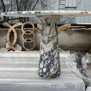 May include: A marble table with a rectangular top and a cylindrical base. The table has a white and gray color scheme, with dark gray veining throughout. The table is approximately 30 inches tall.