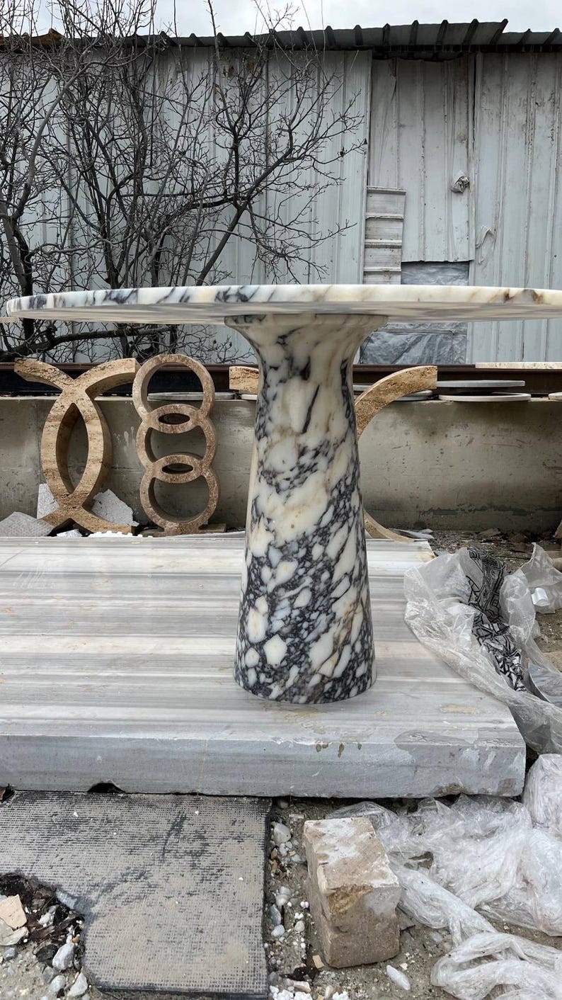 May include: A marble table with a rectangular top and a cylindrical base. The table has a white and gray color scheme, with dark gray veining throughout. The table is approximately 30 inches tall.