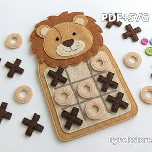 May include: A felt tic-tac-toe game featuring a lion design. The game board is tan with a grid, and the pieces are brown crosses and tan circles. Additional buttons in various colors are scattered around the board. The text "PDF+SVG" and "ByFeltStore" are visible.