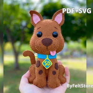 May include: A brown felt dog plush toy with black button eyes, a pink tongue, and dark brown spots. It wears a bright blue collar with a yellow diamond-shaped tag featuring the letters SD. The text PDF+SVG is visible in the upper right corner.