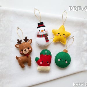May include: Five felt Christmas ornaments featuring a brown reindeer, a snowman with a red and green scarf, a yellow star, a red mitten with a snowflake, and a green bauble. Each ornament has a gold hanging loop and the text PDF+SVG ByFeltStore.