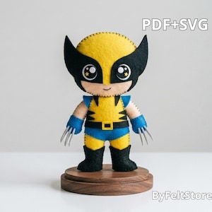 Op de afbeelding: Een viltpop van een personage in een geel en blauw pak met een zwart masker, zwarte laarzen en zilveren klauwen aan elke hand. De pop heeft grote ogen met steraccenten en staat op een ronde houten voet. Tekst leest PDF+SVG.