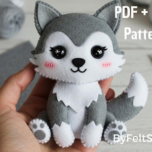 Può includere: Un peluche a forma di lupo fatto a mano in feltro grigio e bianco, con occhi neri a bottone e guance rosa. Il peluche ha un'espressione sorridente e tiene un modello PDF + SVG. Il testo "ByFeltStore" è visibile.