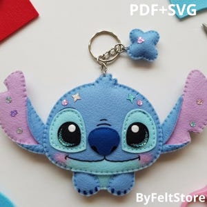 Peut inclure: Un porte-clés Stitch en feutre bleu avec un anneau argenté. Le porte-clés représente le visage de Stitch avec de grands yeux, un nez bleu et des oreilles roses. Il y a aussi un petit charme de fleur bleue. Le texte "PDF+SVG" est visible.