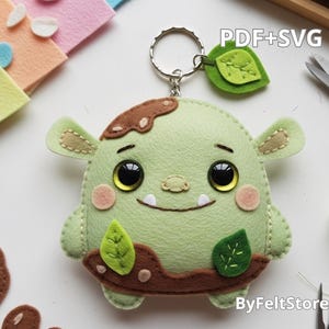 Puede incluir: Un llavero de fieltro hecho a mano que representa un monstruo verde con grandes ojos negros, pequeños dientes blancos y detalles marrones. El llavero incluye un anillo de metal y un dije de hoja verde. El texto "PDF+SVG" es visible.