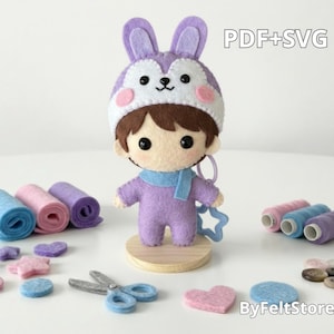 Pode incluir: Boneca de feltro com chapéu de coelho roxo, cachecol e roupa. Rolos de feltro azul, rosa e roxo estão presentes, juntamente com formas de feltro, tesouras e carretéis de linha. O texto "PDF+SVG" é visível.