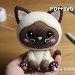 Può includere: Un peluche fatto a mano a forma di gatto siamese con corpo color crema, muso, orecchie e zampe marroni e grandi occhi neri. Il gatto ha guance rosa e baffi. L'immagine include il testo "PDF+SVG" e "ByFeltStore".
