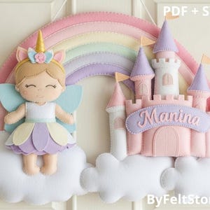 Può includere: Decorazione da parete in feltro con una fata e un castello. La fata ha un corno d'oro, ali blu e un vestito pastello. Il castello è rosa e bianco con torrette viola e il nome "Manina" su uno stendardo. Un arcobaleno sovrasta la scena, su nuvole bianche.