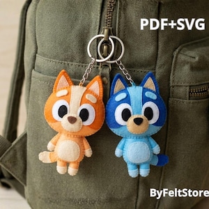 Pode incluir: Dois porta-chaves de feltro em forma de cães de desenho animado, um laranja e um azul, presos a um anel de metal. O cão laranja tem detalhes em creme e o azul tem detalhes em azul claro. O texto PDF+SVG e ByFeltStore é visível.