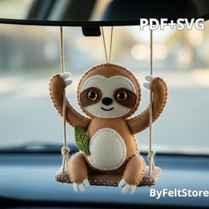 May include: A brown felt sloth ornament hanging from a car mirror. The sloth is on a swing, with a light brown body, a dark brown face, and a green leaf. The ornament is handmade and has the text "PDF+SVG" and "ByFeltStore".