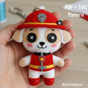 Può includere: Un portachiavi a forma di cane in feltro fatto a mano, rosso, bianco e beige. Il cane indossa un cappello e un gilet da pompiere rossi con una striscia gialla. Gli occhi sono bottoni neri e le guance rosa. Il testo sull'immagine recita "PDF + SVG Pattern" e "ByFeltStore."