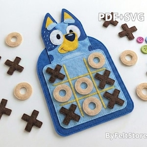 Puede incluir: Un juego de tres en raya de fieltro azul con un diseño de perro de dibujos animados. El juego incluye X de fieltro marrón, O beige y botones de colores. El tablero es azul con líneas amarillas. El texto "PDF+SVG" y "ByFeltStore" son visibles.