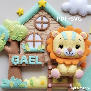 May include: A felt lion cub sits on a wooden structure with a house and tree design. The lion is yellow and orange, with a light blue stripe on its head. The name "GAEL" is displayed on a sign. The image includes the text "PDF+SVG" and "ByFeltStore".