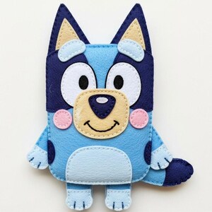 May include: A handmade felt Bluey plush toy. The toy features a light blue body with darker blue accents, including ears and spots. It has a tan nose, white eyes, and pink cheeks. The plush is designed to resemble the cartoon character Bluey.