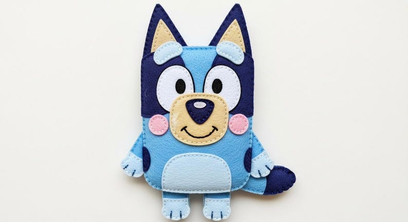May include: A handmade felt Bluey plush toy. The toy features a light blue body with darker blue accents, including ears and spots. It has a tan nose, white eyes, and pink cheeks. The plush is designed to resemble the cartoon character Bluey.