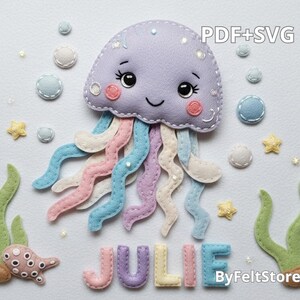 May include: A felt jellyfish with a smiling face, pastel tentacles, and the name "JULIE" in colorful letters. The design includes bubbles, starfish, and seaweed, with the text "PDF+SVG" and "ByFeltStore".