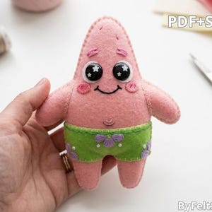 May include: A pink felt plush toy shaped like a starfish with black eyes featuring white star accents, pink embroidered cheeks, and a smiling mouth. The toy wears green felt shorts with purple flower embroidery and white stitching. Text reads PDF+SVG.