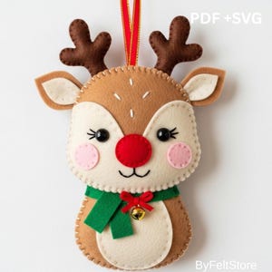 Puede incluir: Adorno navideño de fieltro con forma de reno, con nariz roja, cuernos marrones y bufanda verde. Tiene una cinta roja para colgar. El texto "PDF + SVG" es visible. El adorno es de ByFeltStore.