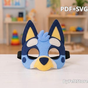 May include: A felt mask shaped like a blue dog with pointed ears, yellow inner ear details, and a black nose. The mask features light blue, dark blue, and cream-colored felt layers with a black elastic strap. Text reads PDF+SVG and ByFeltStore.