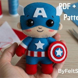 Pode incluir: Boneco de feltro do Capitão América com um traje azul e branco, um escudo e a letra "A" na máscara. O boneco é segurado em uma mão. O texto na imagem diz "PDF + SVG Pattern" e "ByFeltStore".